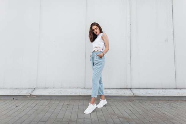 写真 白いスニーカーでヴィンテージの青いズボンのファッショナブルなレースのtシャツを着たモダンな美しい若い女性は、街の壁の近くに立っています。
