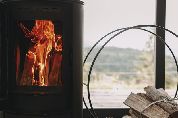 山の景色を望む窓の金属スタンドに火と薪を置いたモダンな黒い暖炉