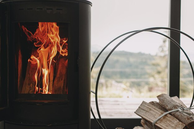 Photo modern black fireplace with fire and firewood on metal stand at window with view on mountains