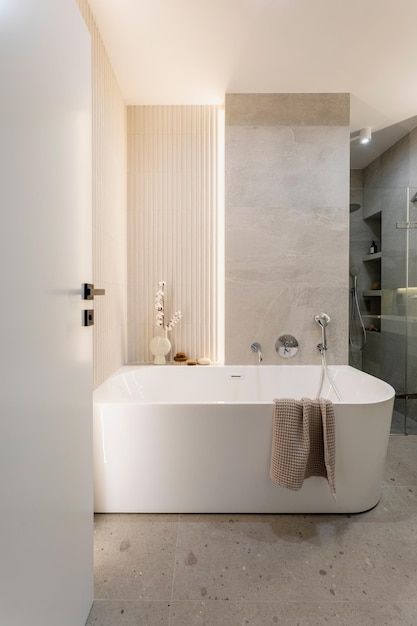 Современная светлая ванная комната с ламельной стеной. Большая белая ванна с серебряным краном и коричневым полотенцем.