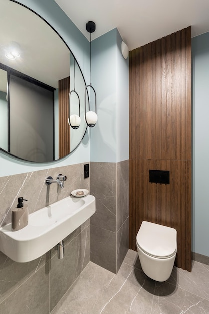 Modern bright bathroom with white toilet seat on wooden lamela wall Round mirror with black frame