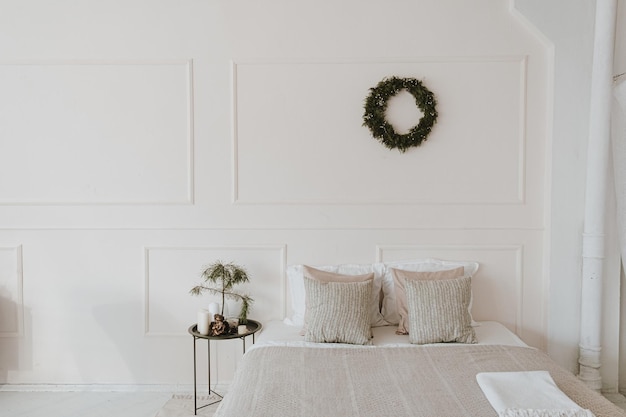 Современный яркий домашний интерьер спальни с кроватью нейтральное постельное белье подушки Рождественский венок из сосновых иголок висит на белой стене Эстетическая гостиная с рождественскими украшениями