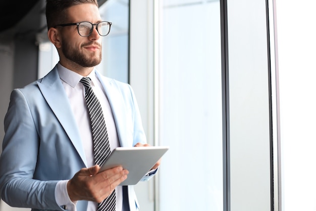 Modern business man in formalwear using digital tablet while standing near window in the office.