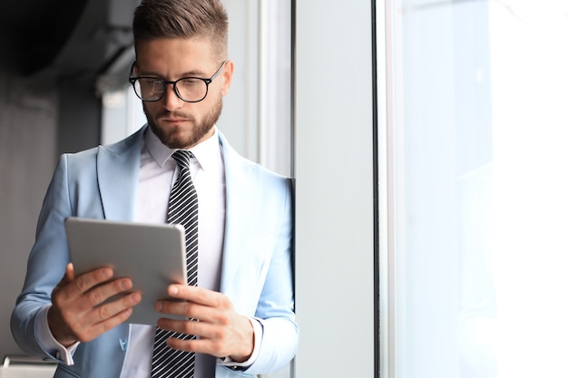 Foto uomo d'affari moderno in abiti da cerimonia che utilizza tablet digitale mentre si trova vicino alla finestra in ufficio.