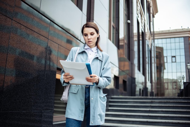 Modern business woman looks attentively into folder with documents on the background of business building with a staircase Lifestyle