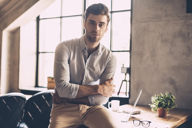 Modern businessman. Confident young man in smart casual wear keeping arms crossed and looking at camera while leaning at the desk in office