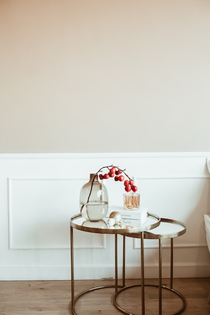 Modern classic interior design. Decorated living room. Bedside table with red berries bouquet in glass vase, book, candle in front of beige wall