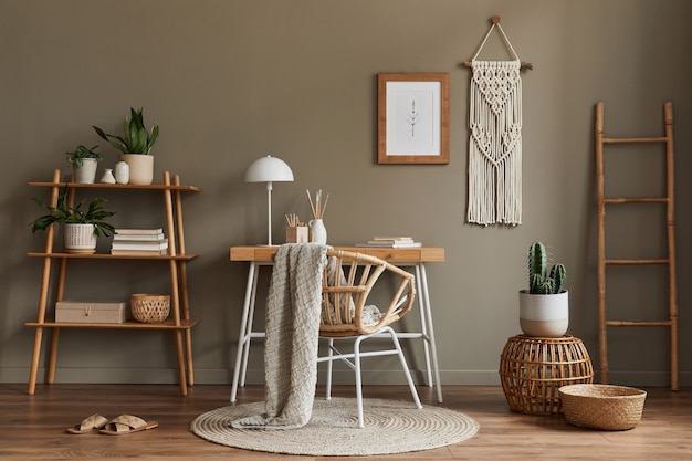 Modern composition at boho interior of home office room with wooden desk, stylish armchair, bamboo shelf, carpet, macrame, frame, office supplies, decoration and personal accessories.