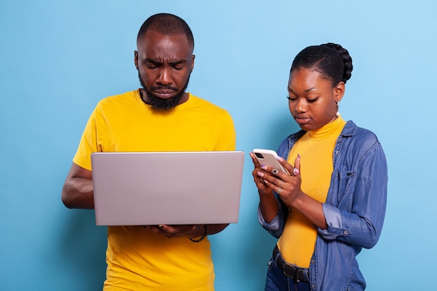 Modern couple browsing internet on laptop and smartphone, using social media application and technology over blue background. Man and woman having fun with computer and mobile phone.