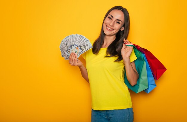 Modern cute smiling young woman with shopping bags and dollars in her hands is posing over yellow wall