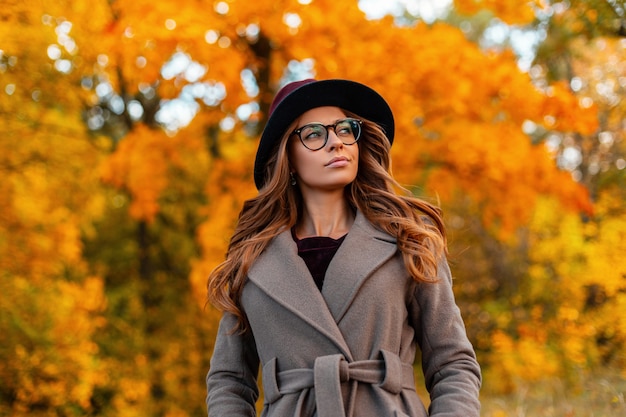 Modern cute young hipster woman with a stylish hairstyle in a vintage hat in fashionable glasses in an elegant coat walks on a the autumn park. trendy girl model enjoys a walk through the woods