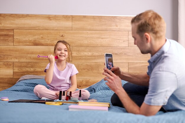 現代のパパは化粧品を使用して彼の流行の娘の写真を撮ります、男と子供は一緒にベッドに座ります
