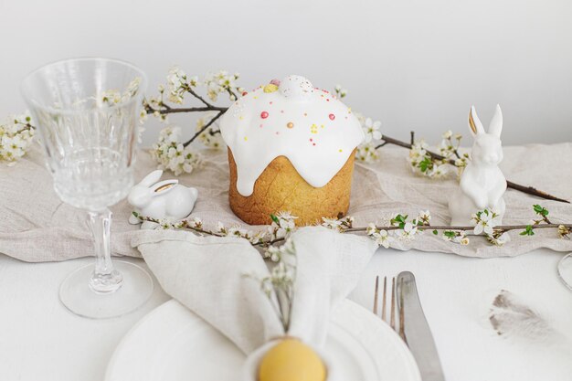 Modern Easter cake egg in bunny napkin on plate cutlery bunny flowers on rustic white table