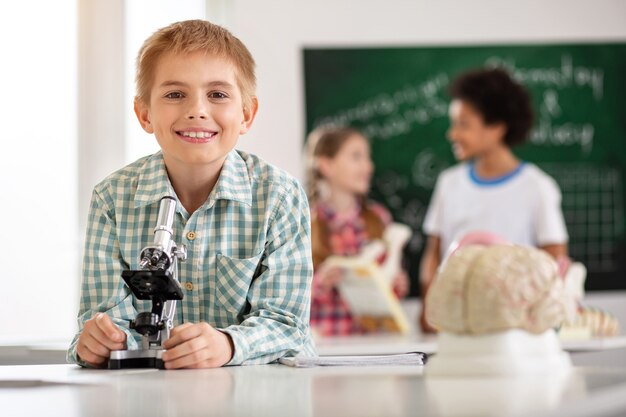 Modern education. Cheerful happy schoolboy standing in front of the microscope while smiling to you