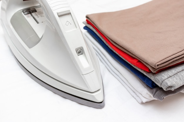 Modern electric iron and a pile of colorful clothes on white background