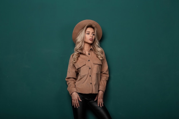 Modern fashionable woman with long blond hair in a beige elegant hat in a vintage shirt in stylish leather pants in retro style posing in a studio near a green wall