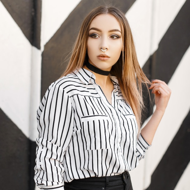 Modern fashionable young woman in a stylish striped shirt with pierced nose in a black velvet necklace in trendy jeans posing near a black and white wall in stripes