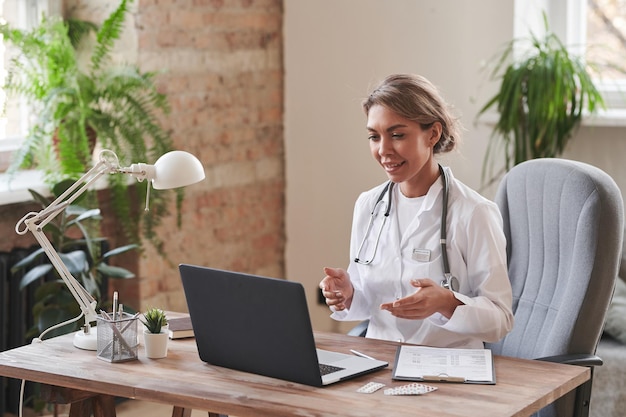 Modern female doctor wearing white coat sitting at desk in loft office talking to patient on video c