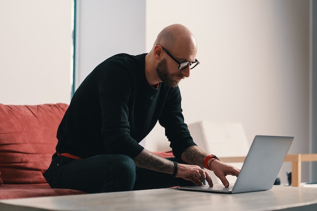 Современный сосредоточенный мужчина с бородой и очками работает на ноутбуке в современном интерьере