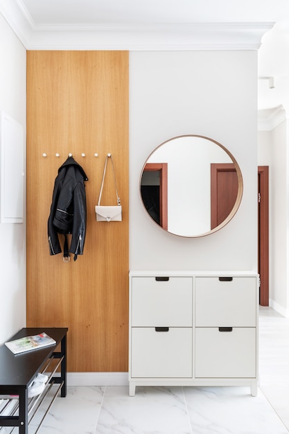 Modern, Fresh White Hallway Interior. Entrance Door, Wooden Hanger With Hanging Clothes And A Lady's Purse. There Are Shoe Cabinet Near The Door, And Round Mirror On The Wall