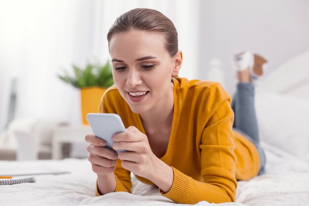 Modern gadget. Pleasant glad pretty woman lying on bed while typing on phone and looking down