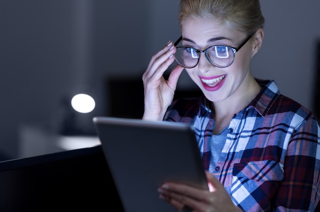 Modern gadget in use. Charming smiling delighted IT girl sitting in the dark lighted room and using modern gadget while expressing positivity and testing the tablet