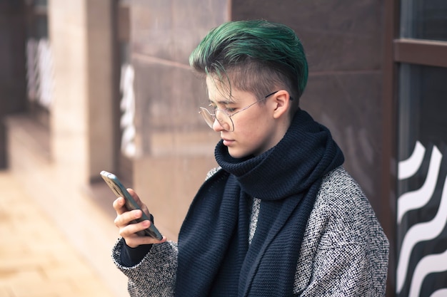 カジュアルな服装の現代の女の子は、秋の街の通りでスマートフォンを使用しています。ソーシャルネットワーク、コミュニケーション。