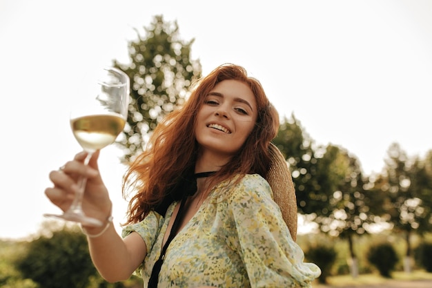 Modern girl with wavy ginger hair in summer yellow clothes and straw hat smiling looking into camera and holding glass with wine outdoor