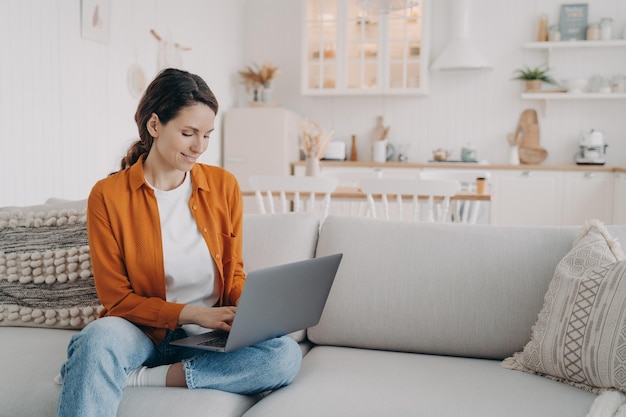 Modern girl working at laptop shopping online chatting on social networks sitting on sofa at home