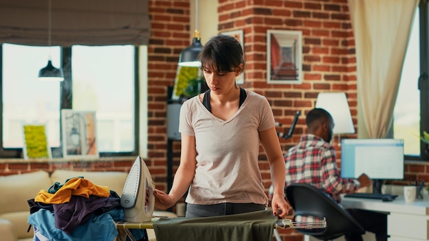 Modern girlfriend using iron with steam to smooth out creases on clothes, ironing pile of laundered garments on ironing board. Casual woman doing spring cleaning at home, housekeeping work.