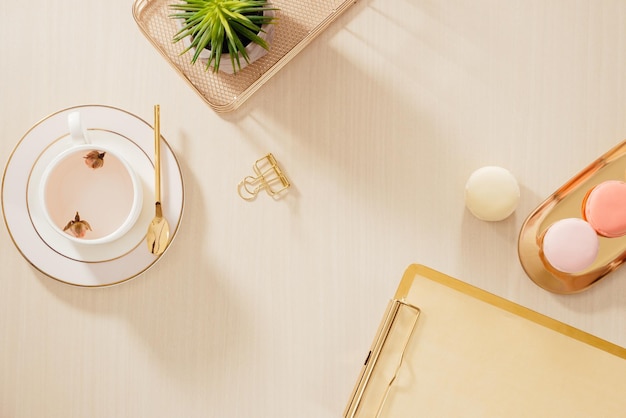 Modern gold stylized home office desk with folder, macaroons, coffee mug on beige background. Flat lay, top view lifestyle concept.`