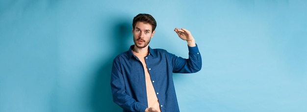Modern guy showing big size shaping large object and looking impressed at camera standing on blue ba