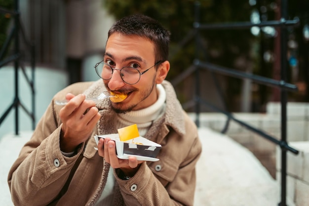 Un ragazzo moderno che indossa gli occhiali seduto nel parco e mangia poffertjes freschi