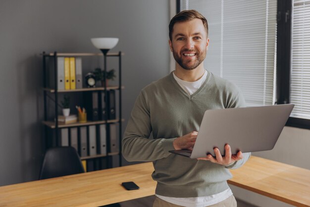 사진 현대적인 행복한 수염이 있는 it 회사의 남성 사무실 직원이 사무실에서 미소 짓고 노트북을 들고 있습니다.