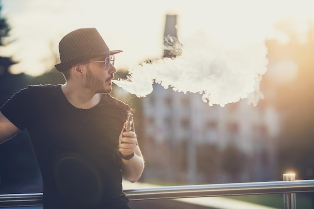 Modern happy young man in hat with a beard fun Vaporizers In the background the evening sunset