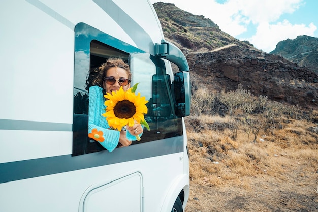 Photo modern hippy travel lifestyle happy young woman out of the camper van window enjoy trip destination and holding big sunflower concept of rent rv vehicle motor home living off grid alternative