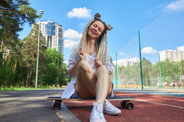 Una moderna ragazza hipster si siede su un longboard sorride e mostra la sua lingua guardando dritto verso la fotocamera fotografia sportiva foto di alta qualità