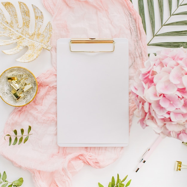 Modern home office desk workspace with blank paper clipboard, pink hydrangea flowers and accessories on white surface