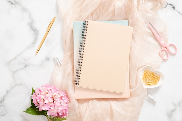 Foto moderna area di lavoro per ufficio a casa con fiori di ortensia rosa, coperta pastello, blocco note di carta bianca, articoli di cancelleria dorati e accessori femminili