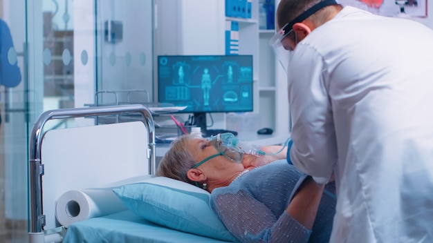 Nel moderno ospedale o clinica, il medico sta mettendo la maschera di ossigeno sul paziente anziano che giace a letto. tema sanitario medicinale correlato al coronavirus covid-19. trattamento delle infezioni durante l'epidemia