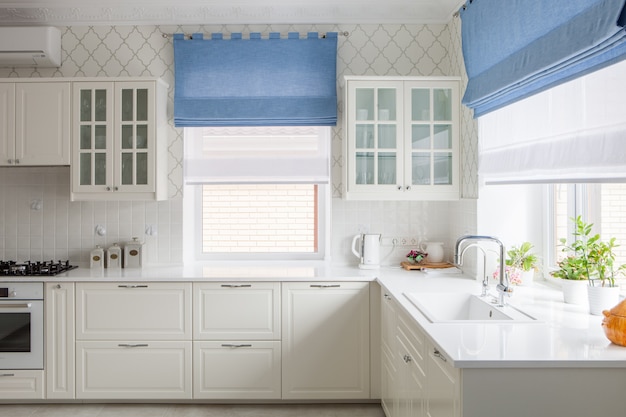 Modern house Interior of spacious bright kitchen with white furniture. Window blue curtains behind the sink