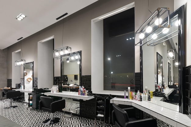 Modern interior of the barbershop with mirrors chairs and other equipment near large windows in the evening