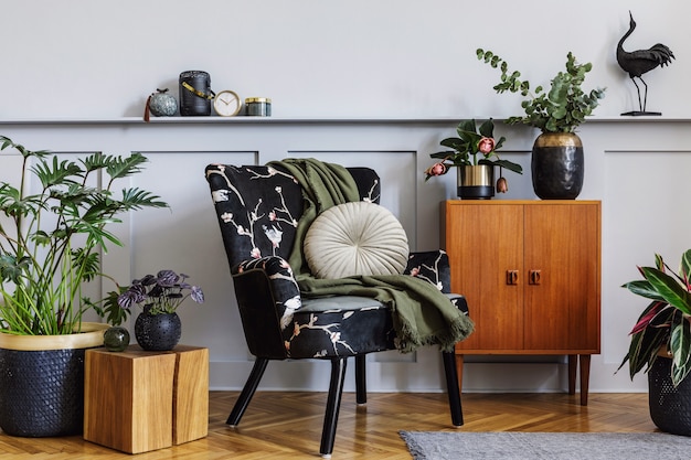 Modern interior of living room with design armchair, wooden\
vintage commode, wood paneling with shelf, a lot of plants, cube,\
carpet, grey wall and persnoal accessories in home decor.