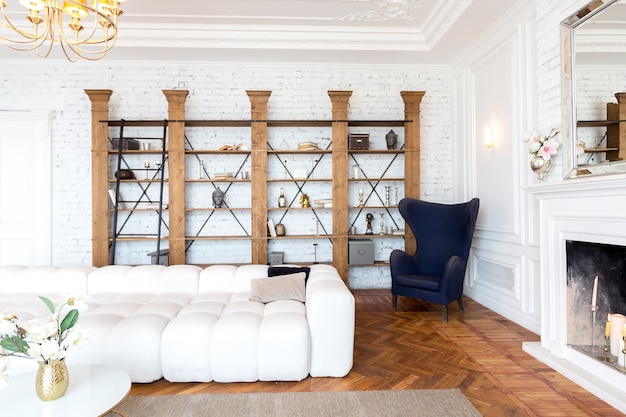 Modern interior of a luxurious large bright living room. white expensive sofa and wooden shelving, white walls with moldings and a luxurious chandelier