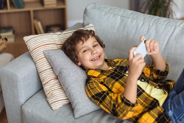 Modern kids leisure Cute boy playing games on mobile phone resting on sofa at home smiling to camera empty space