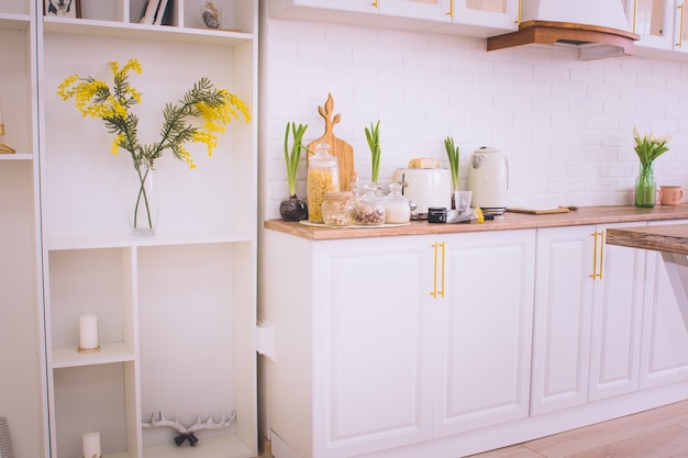 Modern kitchen interior with white facades, mimosa flowers, kettle, cans of groceries