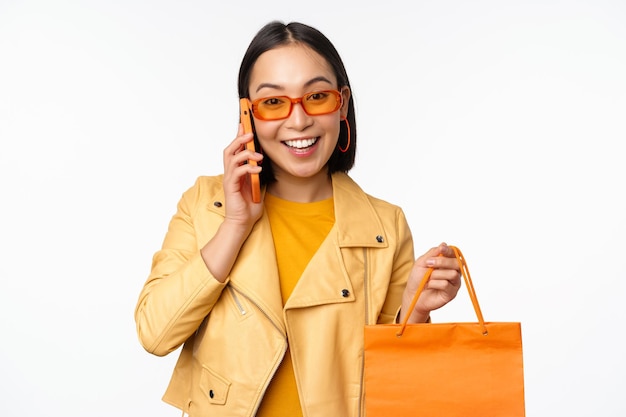 Modern korean girl on shopping holding store bag talking on mobile phone and smiling standing over white background