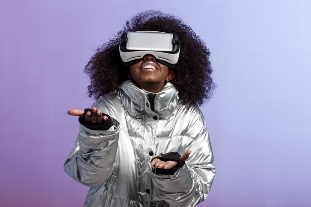 Modern krullend bruinharig meisje gekleed in een zilverkleurige jas maakt gebruik van de virtual reality-bril poses in de studio op neon achtergrond.