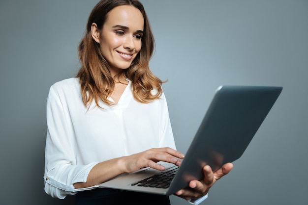 Modern laptop. Joyful nice happy woman holding a laptop and looking at its screen while pressing a button