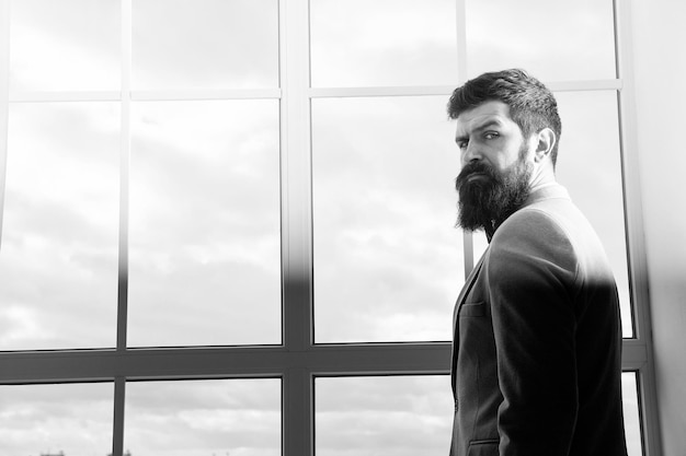 Modern life business man standing at window with cityscape Brutal bearded man Mature hipster with beard Male in modern business office Businessman in formal suit Modern life of businessman